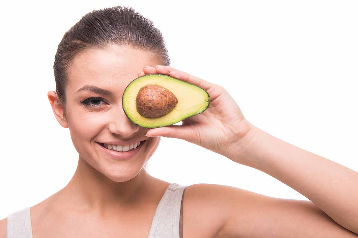 A Smiling Woman Holding an Avocado Half To Her Eye.