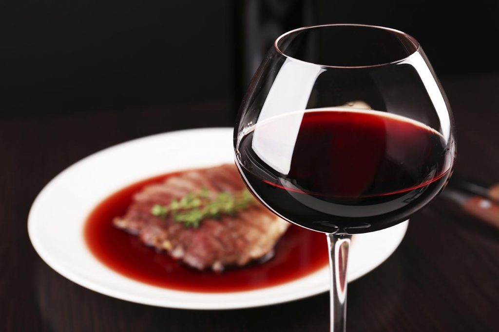 A glass of red wine next to a plate of food on the table.
