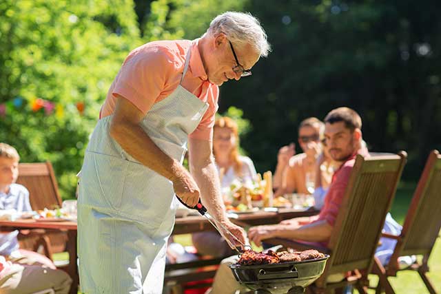 An Elderly Man Cooking Meat On a BBQ Grill.