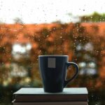 A cup on top of a book in front of a rain-splashed window.