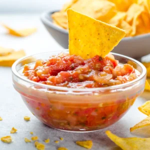 A small glass bowl of salsa with a tortilla chip in it.