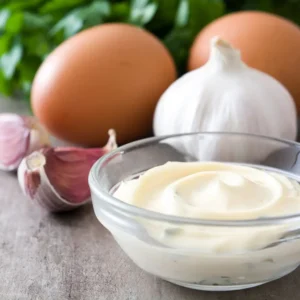 A small glass bowl of aoili sauce next to garlic and two eggs.