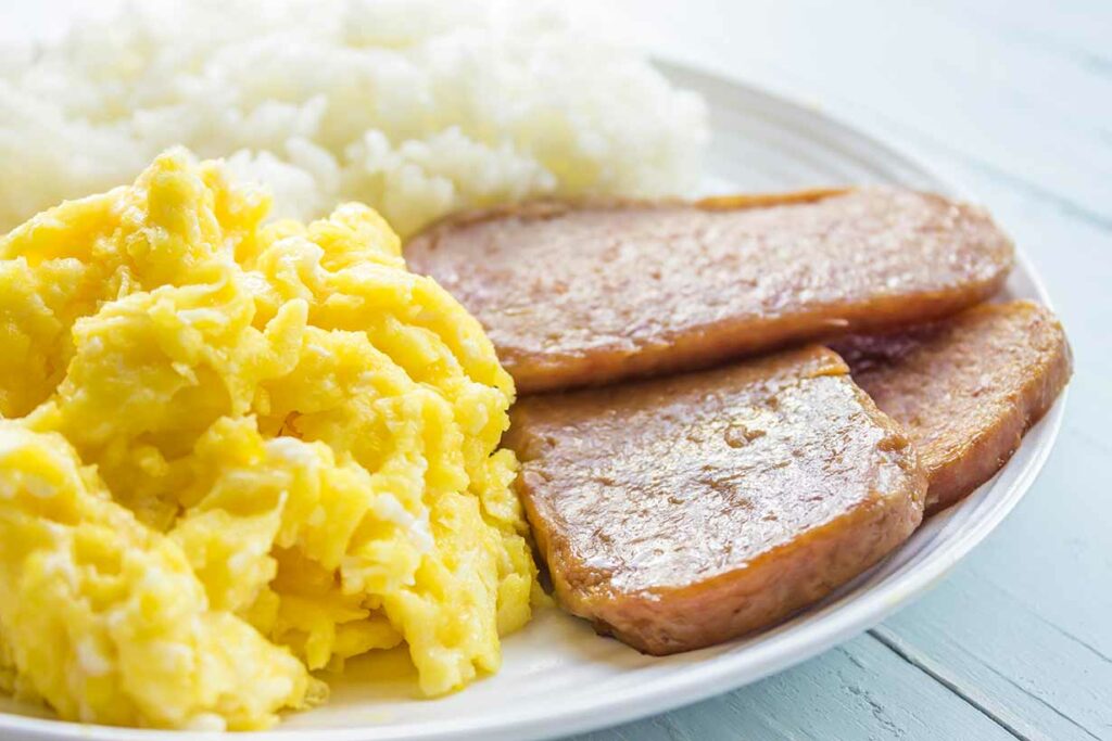 Cooked slices of spam on a plate alongside other foods.