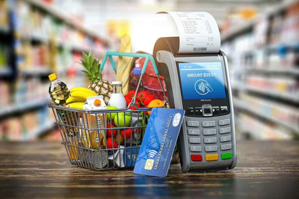 A Shopping Basket Filled With Foods Next To a Payment Machine Displaying Cost (Food Budgeting Theme).