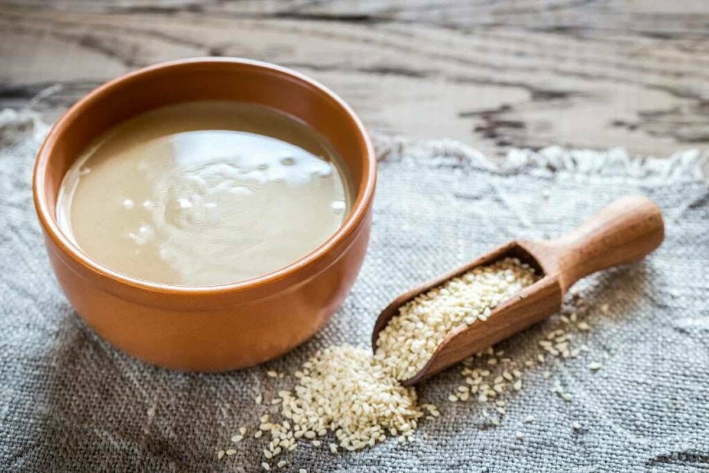 A bowl of tahini next to sesame seeds.