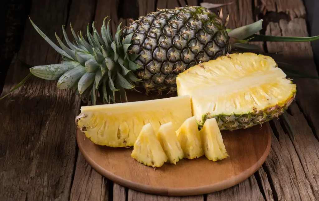 A whole pineapple next to various sizes of chopped pineapple on a wooden board.