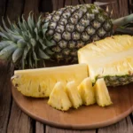 A whole pineapple next to various sizes of chopped pineapple on a wooden board.