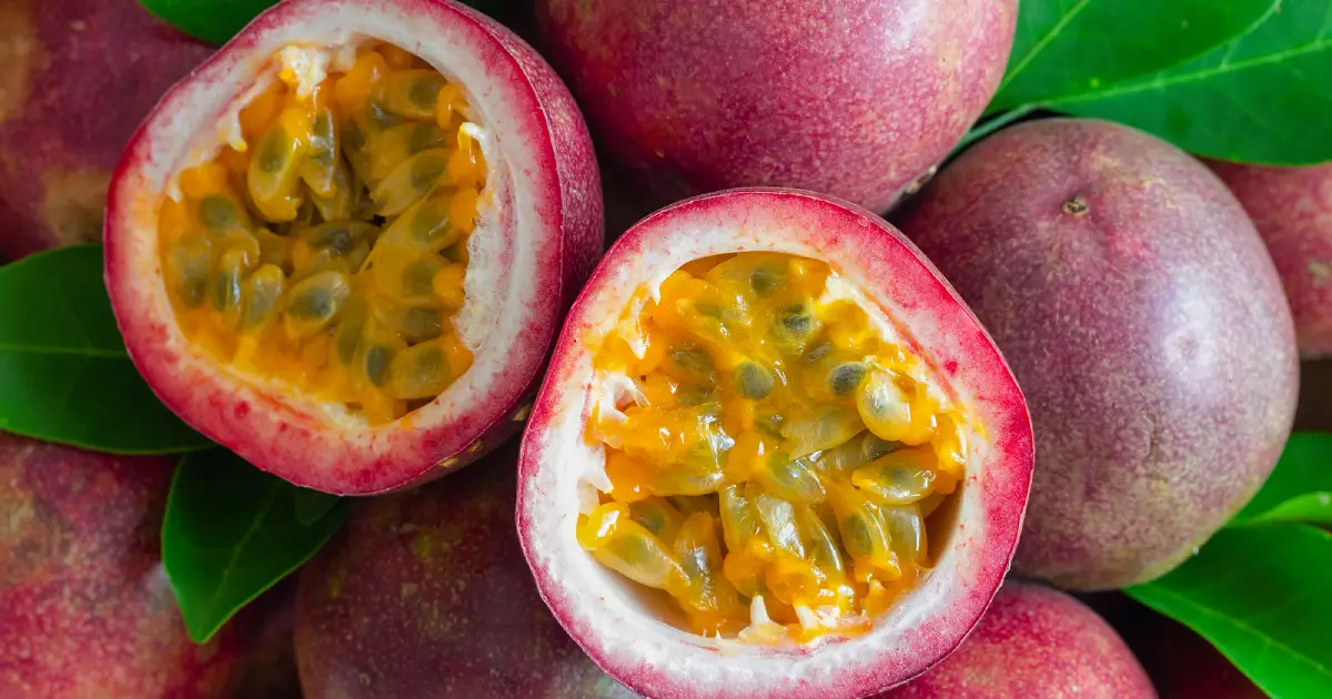 Whole purple passion fruits next to a passion fruit cut into two halves showing the inner yellow pulp.