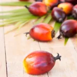 Palm Oil: Nutrition Profile and Health Effects, According to Science Glass bottle of palm oil on a wooden surface next to palm fruits.