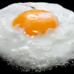 30 Common Foods High in Dietary Cholesterol A runny fried egg frying on a black surface.